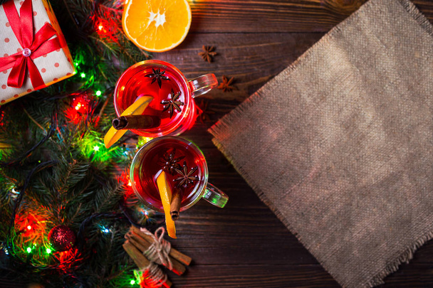 Two glasses,cup of red hot drink.Mulled wine on wooden background with christmas spices,orange slice,close up.Christmas mulled wine.Branch of Christmas tree, Christmas lights - 写真・画像