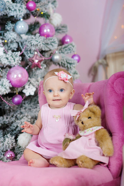 charming little girl with blue eyes sitting in a pink armchair teddy bear on background decorated Christmas tree - Photo, Image