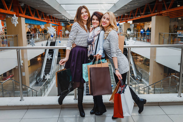 τρεις όμορφες νεαρές γυναίκες με τσάντες αγορών βλέπουν φωτογραφική μηχανή στο εμπορικό κέντρο - Φωτογραφία, εικόνα