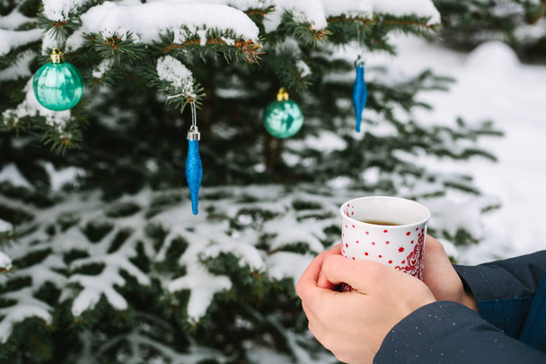 man holding a cup of hot tea in his hands next to the Christmas tree in winter forest in the New Year holidays - Photo, Image