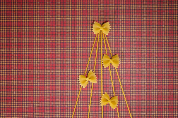 Christmas tree silhouette made of spaghetti and bow tie pasta on red squared background. Flat lay. Top view. Winter holidays concept - Photo, Image