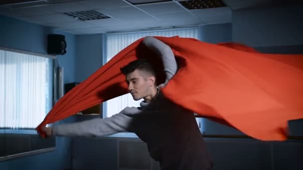 Close up of handsome male dancer in grey pullover and black trousers dancing in the classroom with red cloth. Young student of dance school is performing element of classical choreography - Imágenes, Vídeo