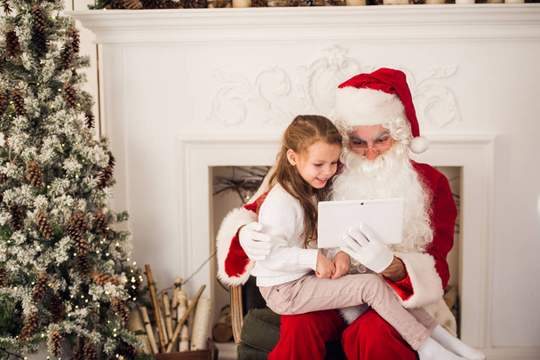 Різдвяні свята щаслива дівчина Санта дивиться щось на цифровому сенсорному екрані планшетного ПК, над димоходом і деревом барвисті вогні фону
. - Фото, зображення