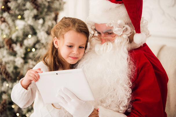 Little girl sitting with Santa using tablet on the armchair at home living room - Photo, Image