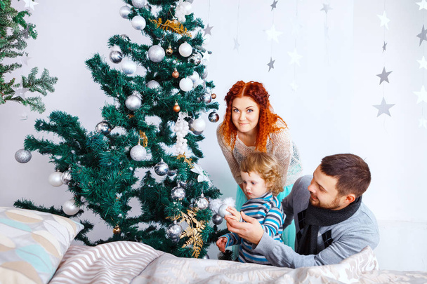 Navidad feliz familia de tres personas decorando abeto con cajas de regalo sobre fondo de dormitorio blanco
 - Foto, imagen