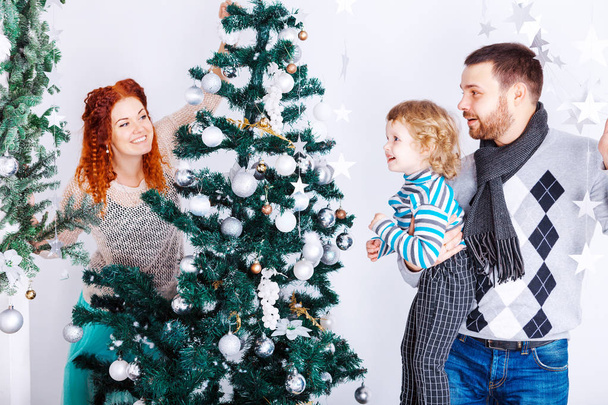 Navidad feliz familia de tres personas decorando abeto con cajas de regalo sobre fondo de dormitorio blanco
 - Foto, Imagen
