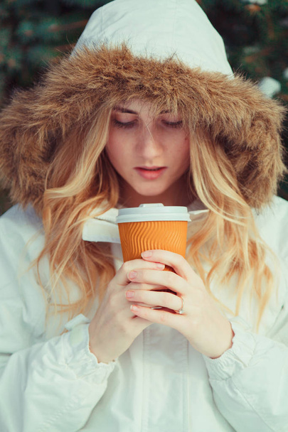 コーヒーのマグカップを保持している毛皮のフードの若い金髪の女性  - 写真・画像