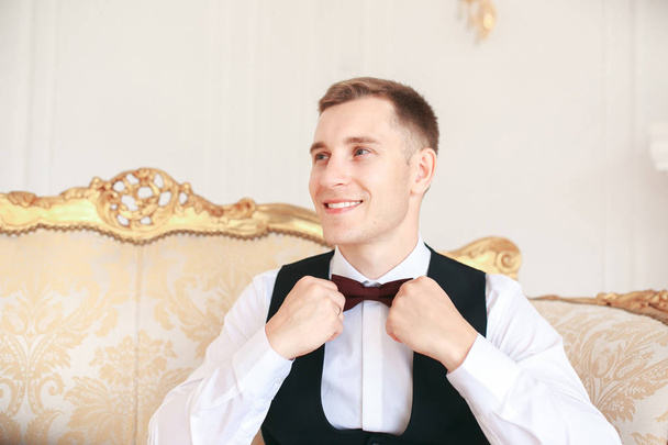  Groom sitting on the sofa waiting for the bride on his wedding day. at wedding tuxedo smiling and waiting for bride.Elegant man in black costume and bow-tie.  - Photo, image