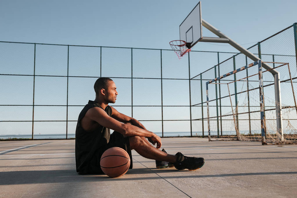 Tired african sports man sitting with basketball at the playground - Фото, изображение