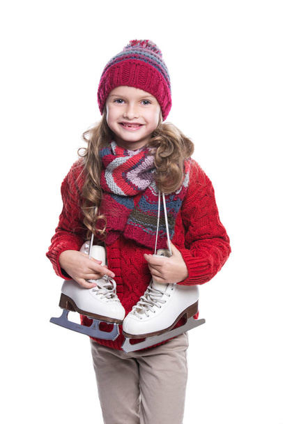 Cute smiling little girl with curly hairstyle wearing knitted sweater, scarf, hat and gloves with skates isolated on white background. Winter clothes and sport concept. - Foto, Bild