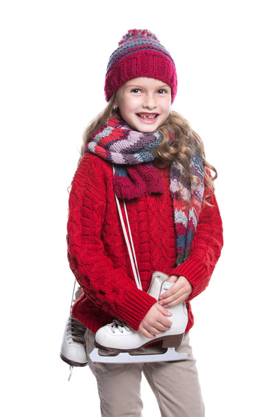 Cute smiling little girl with curly hairstyle wearing knitted sweater, scarf, hat and gloves with skates isolated on white background. Winter clothes and sport concept. - Zdjęcie, obraz
