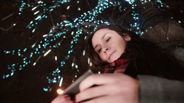 young attractive woman using smartphone at snowy Christmas night standing under a tree decorated with sparkling lights - Photo, Image