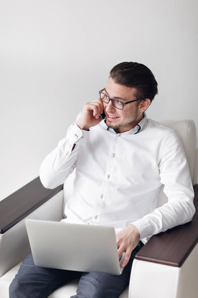 Portré, fiatal ember a szemüveg használata mobiltelefon ül míg Laptop szék. Fehér háttér - Fotó, kép