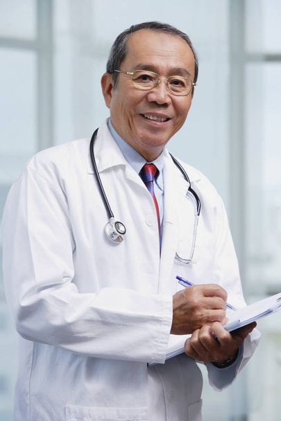 Doctor writing on medical chart, looking at camera - 写真・画像