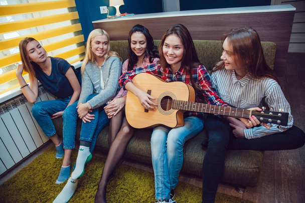 Beautiful girls sitting on couch, have fun and play the guitar in Hostel. - Foto, afbeelding