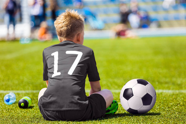 Niño en ropa deportiva negra con pelota de fútbol. Niño sentado en el campo de fútbol. Partido de fútbol escolar para niños
 - Foto, imagen