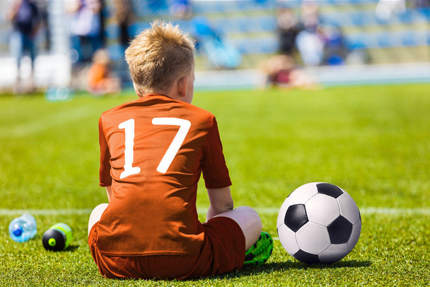 Młody piłkarz Football. Mały chłopiec siedzi na boisko do piłki nożnej. Piłkarz młodzieżowej w Orange Soccer Jersey - Zdjęcie, obraz