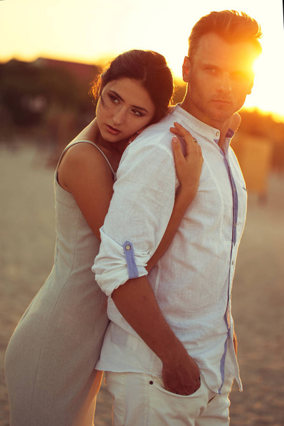 fashionable young couple standing, resting on the beach, wearing in a white clothes posing love at sunset. with custom white balance, color filters, and some fine film noise added to the image - Фото, зображення
