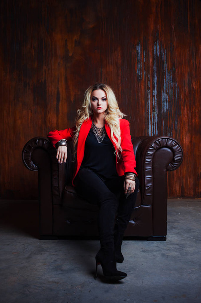 Mulher loira jovem e atraente em jaqueta vermelha senta-se em poltrona de couro, fundo grunge parede enferrujada
 - Foto, Imagem