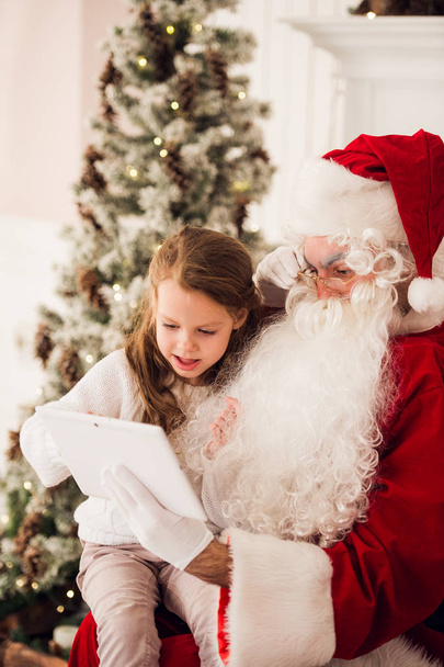 Little girl sitting with Santa using tablet on the armchair at home living room - Photo, image