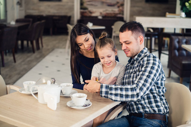 Familie, Elternschaft, Technologie-People-Konzept - glückliche Mutter, Vater und kleines Mädchen beim Abendessen mit dem Smartphone im Restaurant - Foto, Bild