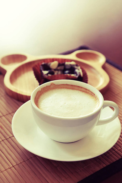 Tasse Kaffee auf Holztisch mit unscharfem Teller von Brownie im Hintergrund.  - Foto, Bild