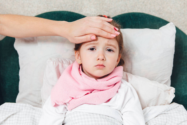 Больной ребенок лежит в постели и трогает ее за лоб.
 - Фото, изображение