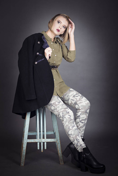 Jovem mulher atraente em roupas de estilo militar sentado no banquinho e segurando um grande casaco na mão
 - Foto, Imagem
