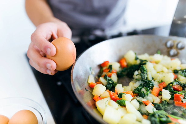 Hands of a young woman adding egg to vegetables into the pan. - Photo, image