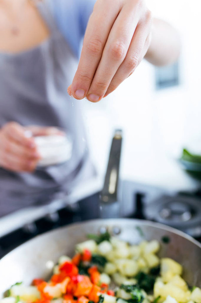 Hand of a young woman adding salt to vegetables into the pan. - Foto, Imagem