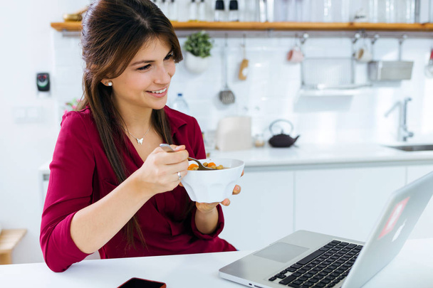 Beautiful young woman working with laptop while eating in the kitchen. - Фото, зображення