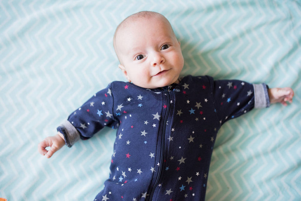baby boy plays on blue blanket, wearing blue sleeper smiling up at camera - 写真・画像