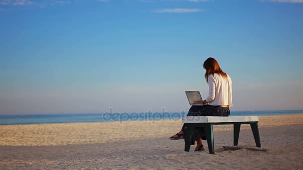 Кавказская девушка пишет текст на ноутбуке, сидя на скамейке на пляже, пузырьки вдоль побережья
 - Кадры, видео