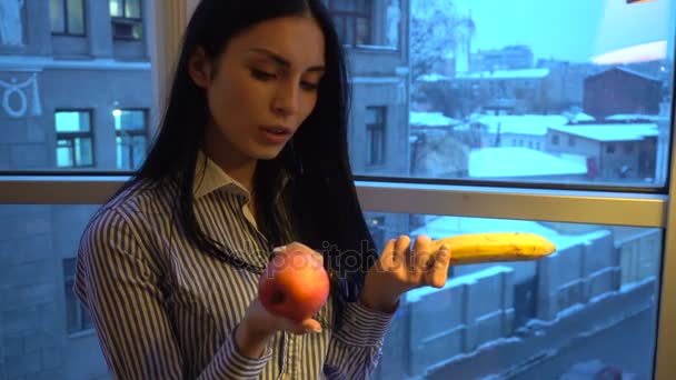 lovely young girl with dark hair are choosing between an Apple and a banana - Imágenes, Vídeo
