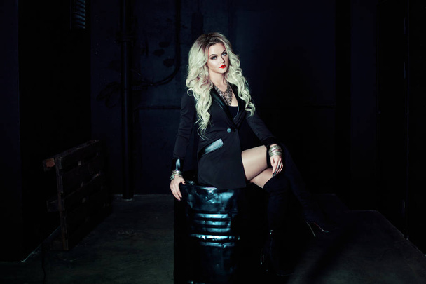 Beautiful fashion blonde woman in black sitting on a metal barrel. Totally dark style - Foto, afbeelding