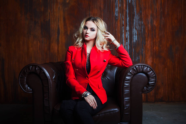 Young and attractive blond woman in red jacket sits in leather armchair, background grunge rusty wall - Foto, Bild