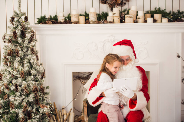 Vánoční dovolená šťastná dívka santa při pohledu na něco na digitální dotykový tablet Pc, přes komín a strom barevné osvětlení pozadí. - Fotografie, Obrázek