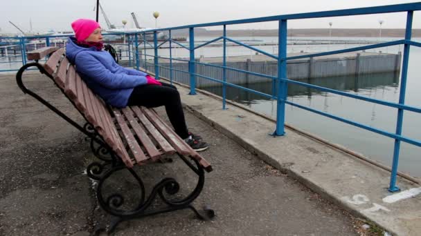 Chica sentada en un banco en el muelle en un día frío y nublado mira a la distancia
 - Imágenes, Vídeo