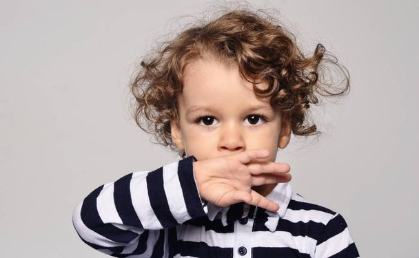 Beautiful curly toddler looking forward and holding his hand on his mouth, boy trying not to speak, shut up - Фото, зображення