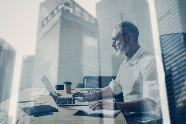 Double exposure concept.Adult bearded businessman working at wood table in modern place.Stylish middle age man using notebook.Skyscraper office building on the blurred background.Horizontal. - Foto, afbeelding