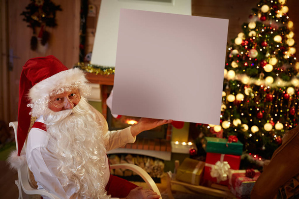 Santa Claus in his living room holding empty white banner  - Fotó, kép