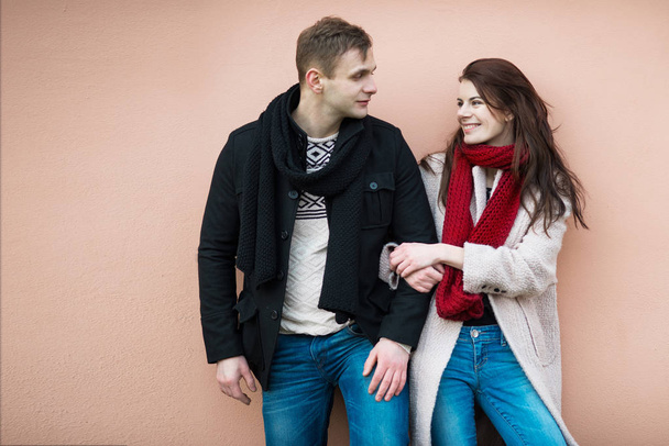 Happy young fashionable couple in love wearing warm stylish winter outfits with jacket, jeans, sweater and scarf standing near the wall and looking on each other smiling. - Photo, image