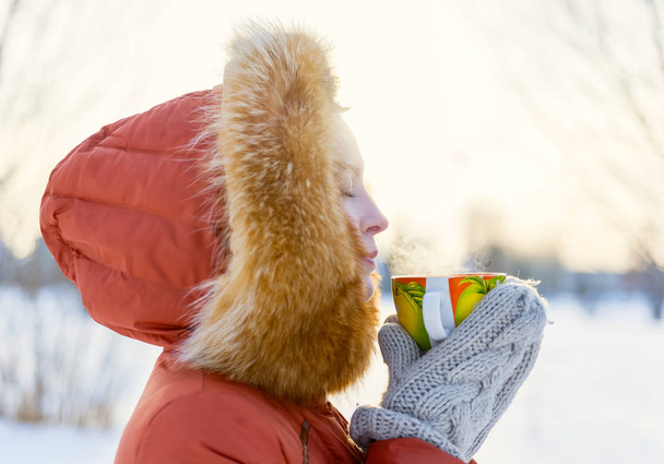 Outdoor portrait of a smiling young girl in a fur hood. Drinking tea from an orange mug in the winter park - Фото, изображение