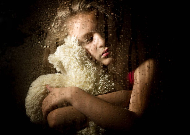 lonely young sad girl behind the window with drops, holding teddy bear and crying - 写真・画像