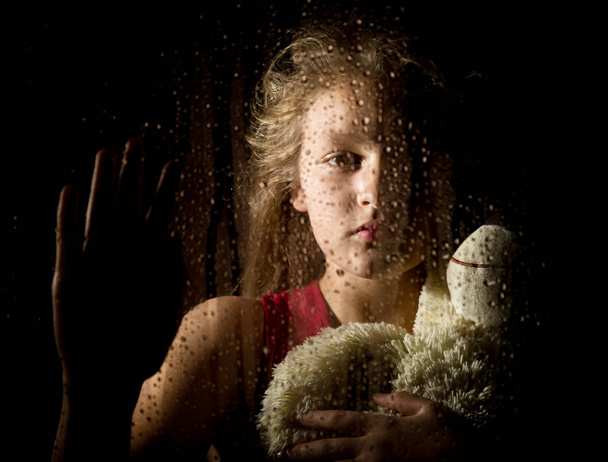 lonely young sad girl behind the window with drops, holding teddy bear and crying - Photo, image