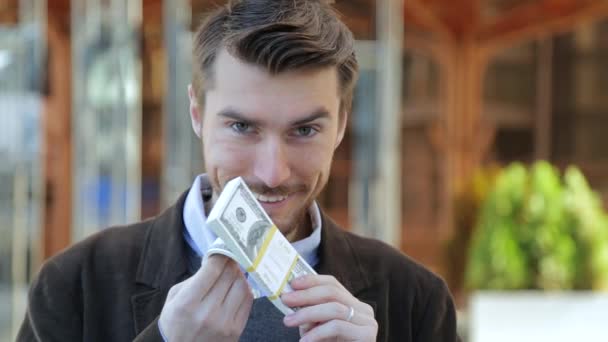 Attractive man leafing through a bundle of dollars, smelling cash, looking at camera and smiling - Imágenes, Vídeo