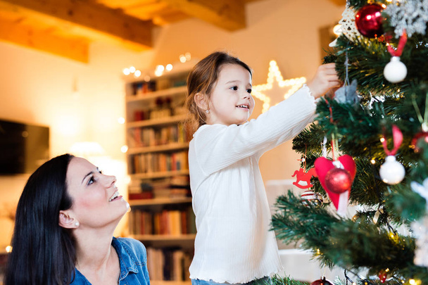 Молодая мама с дочерью украшают елку вместе
. - Фото, изображение