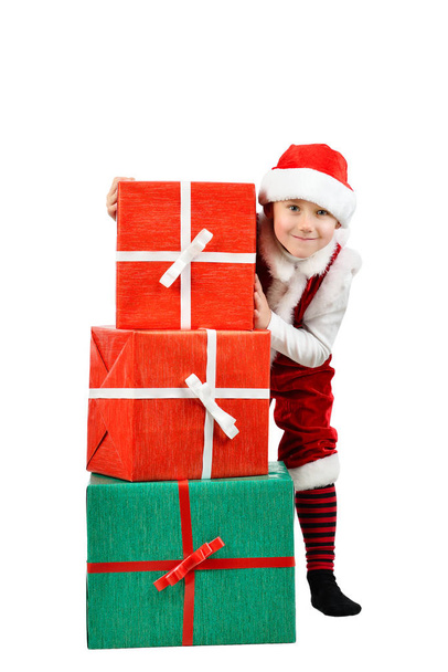 Adorable boy in santa clothes peeks out behind Christmas big gift boxs. Isolated white background. - Zdjęcie, obraz