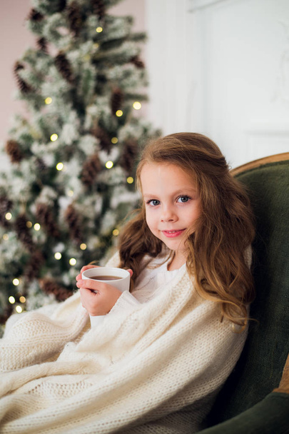 Little girl wearing pajama sitting on armchair wrapped in a blanket with holiday bedding by Christmas tree and fireplace drinking milk at home - Zdjęcie, obraz
