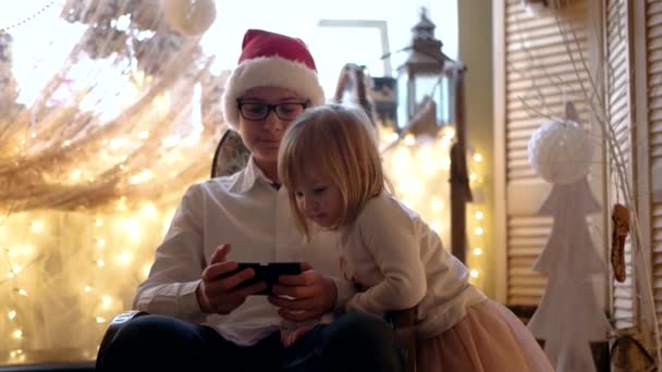 A kis aranyos lány és Santa kalap birtoklás móka-val mozgatható telefon, a hintaszékben idősebb bátyja - Felvétel, videó